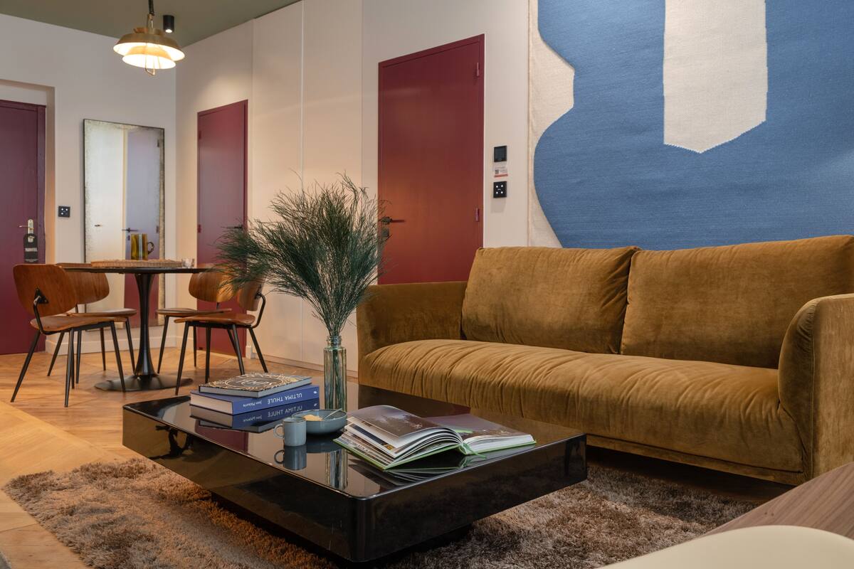 A modern living room with a brown sofa, a glass coffee table with books, a vase with plants, and a large abstract painting on the wall.