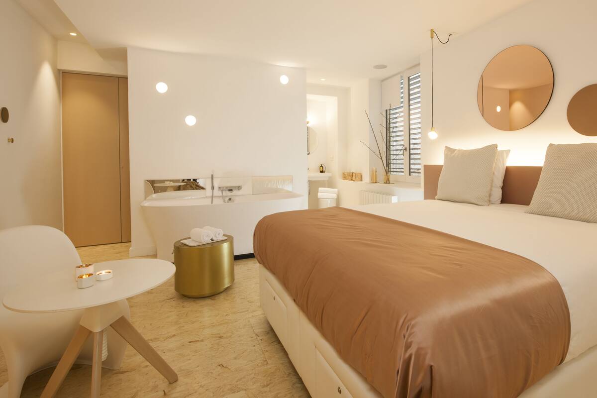 A modern hotel room with a large bed, a round mirror, a small round table, and a separate bathroom area.