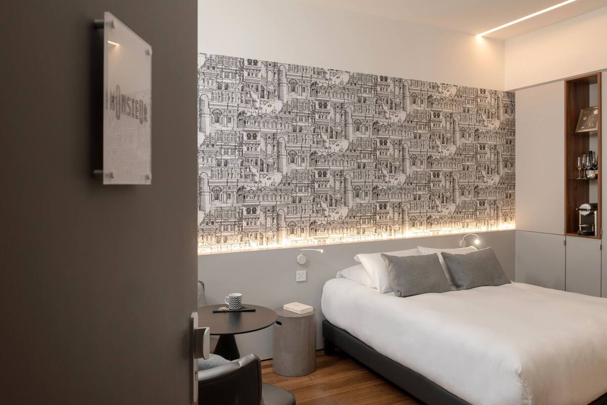 A modern hotel room with a large bed, a small table, and a wall with a detailed cityscape wallpaper.