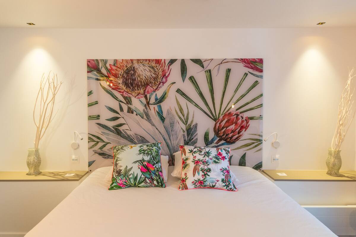 A bedroom with a large floral artwork on the wall, two bedside tables with vases, and a bed with two pillows.