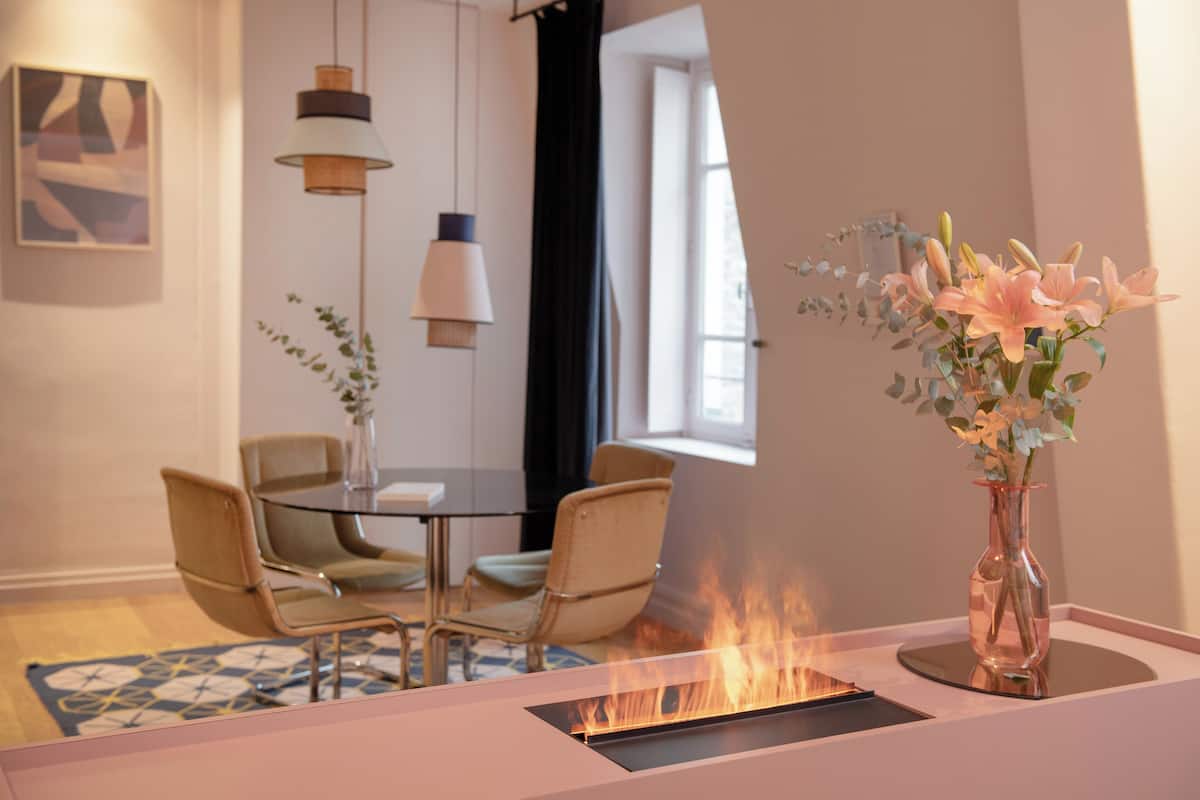 A modern dining area with a glass table, two chairs, a vase with flowers, and a fireplace.