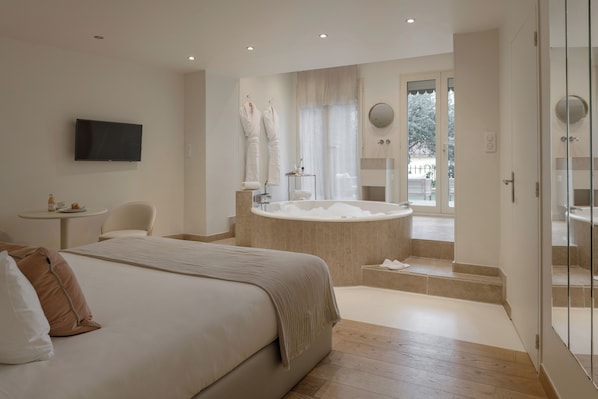 A modern hotel room with a large bed, a sitting area with a table and chairs, a bathtub, and a television mounted on the wall. - MiHotel (Lyon)