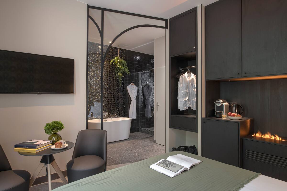 A modern hotel room with a bed, a small table with books, a chair, a TV, a fireplace, and a bathroom with a shower and bathtub.