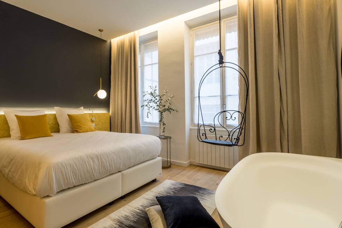 A modern bedroom with a large bed, bedside lamps, a hanging chair, and a bathtub.