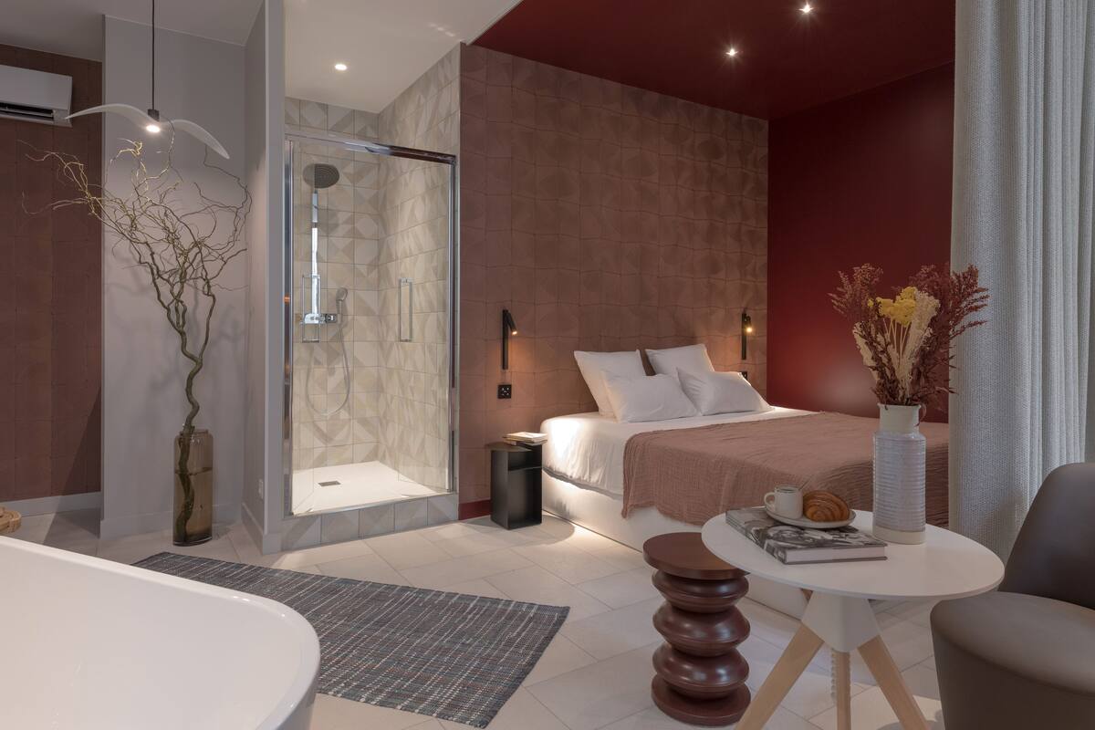 A modern bedroom with a large bed, a bedside table with a book and a cup, a shower area, and a small round table with a vase.
