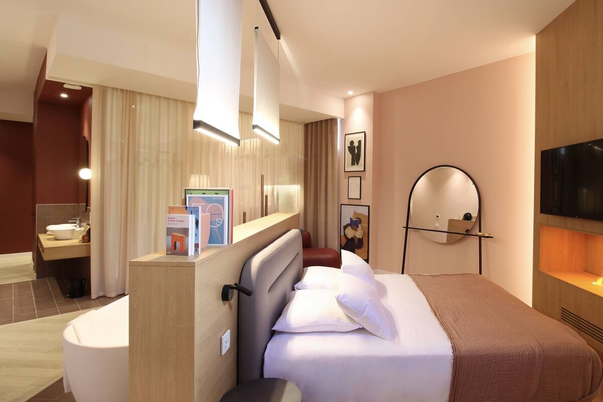 A modern hotel room with a bed, bedside table, TV, fireplace, and a bathroom with a sink and mirror.