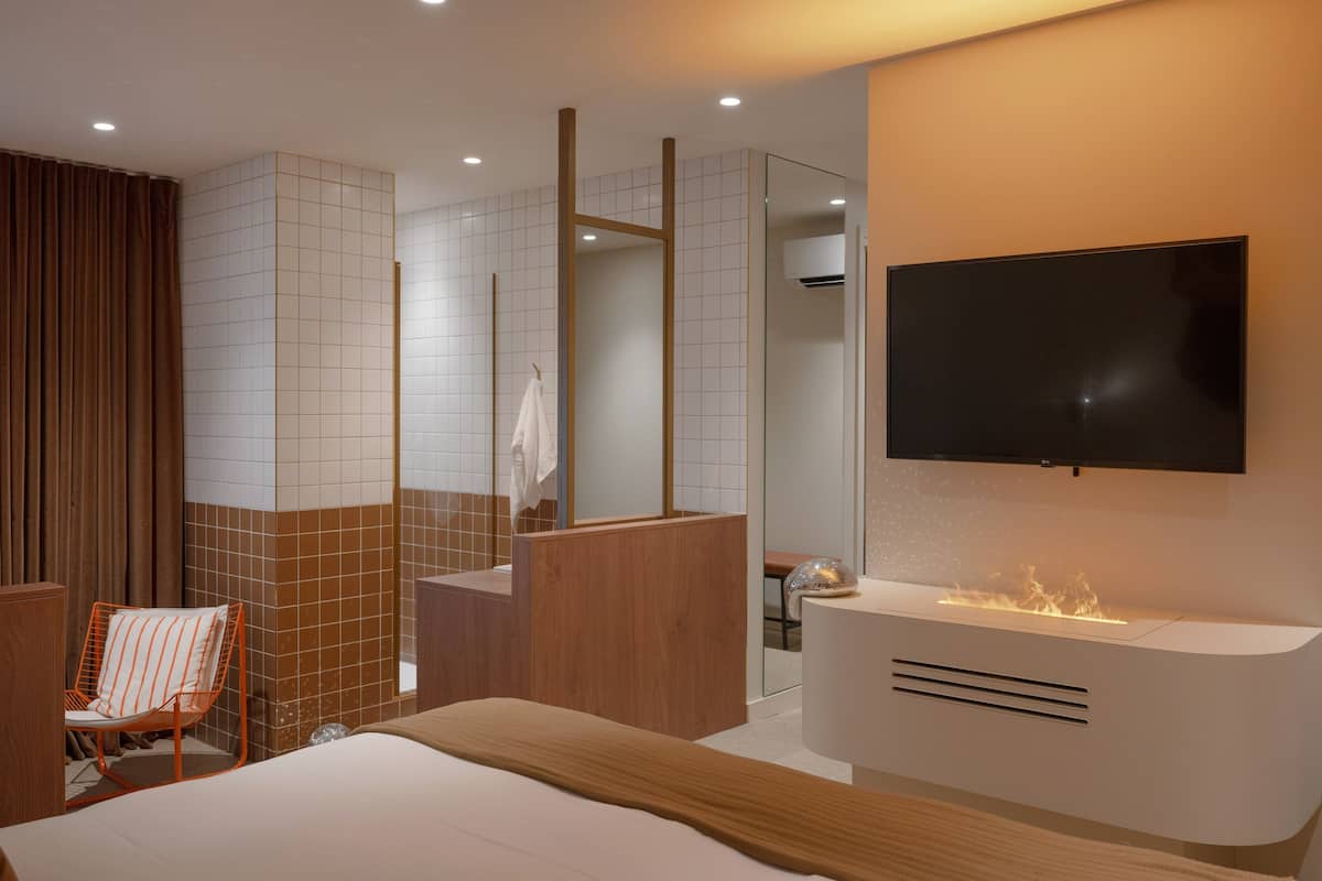 A modern hotel room with a bed, a TV mounted on the wall, a shower area, and a wooden chair.
