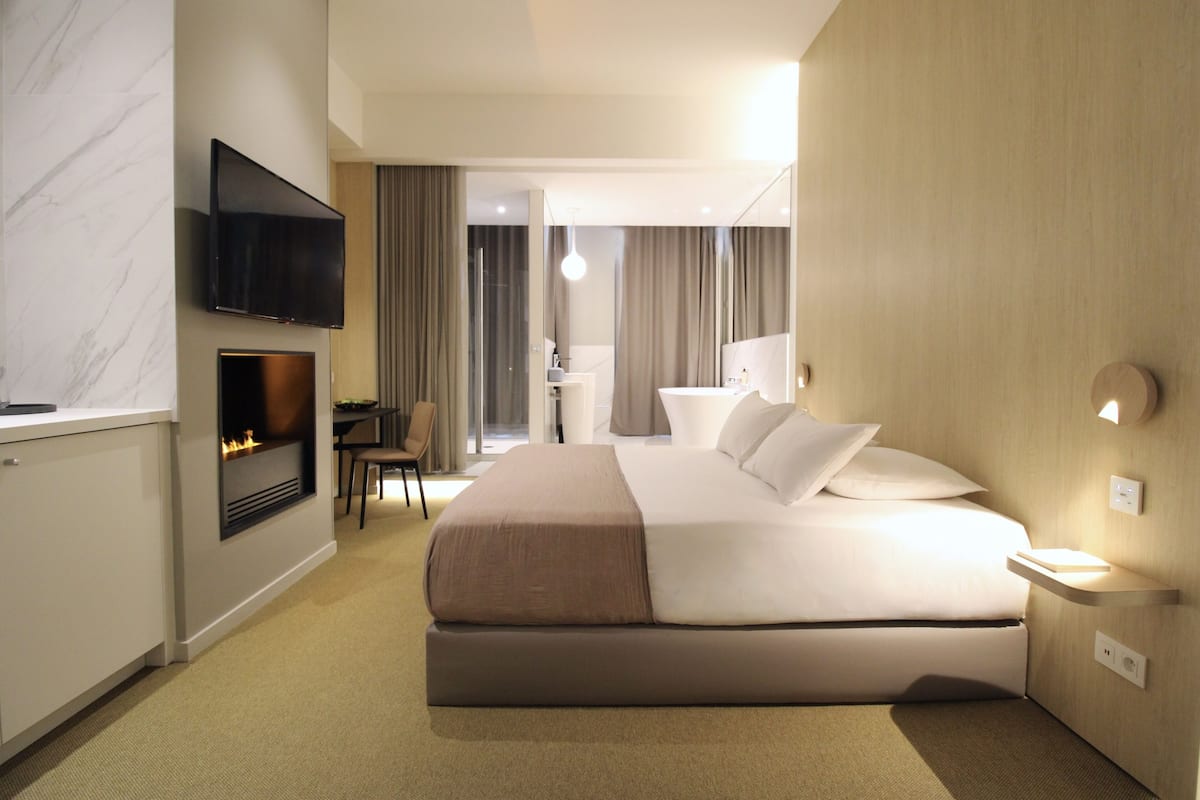 A modern hotel room with a large bed, a flat-screen TV mounted on the wall, a fireplace, a desk, and a bathroom visible through an open door.