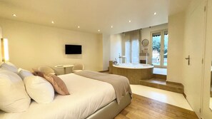 A modern bedroom with a large bed, a small table, a TV, and a bathroom with a bathtub and shower. - MiHotel (Lyon)
