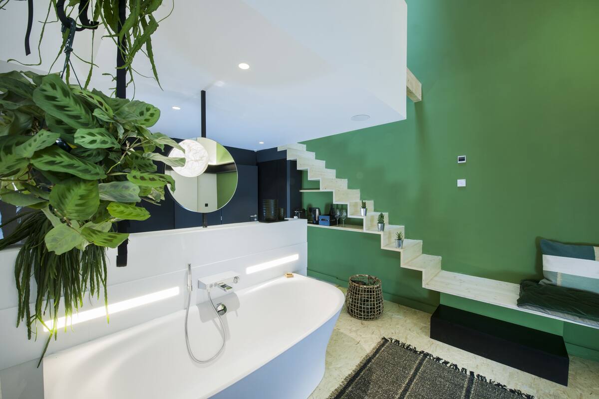 A modern bathroom with a freestanding bathtub, a green accent wall, and a staircase leading to a loft bed area.