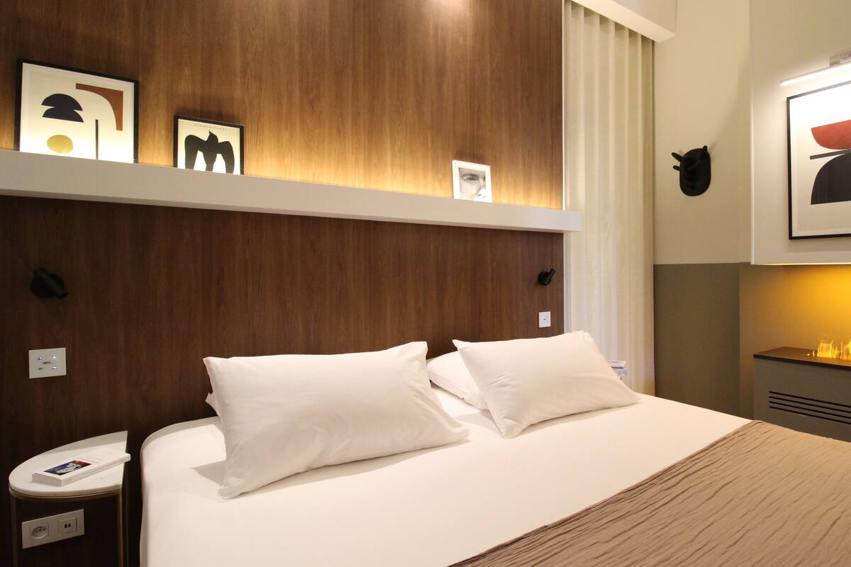 A neatly made bed with white linens and a wooden headboard. There are framed artworks above the bed and a wall-mounted heater to the right.