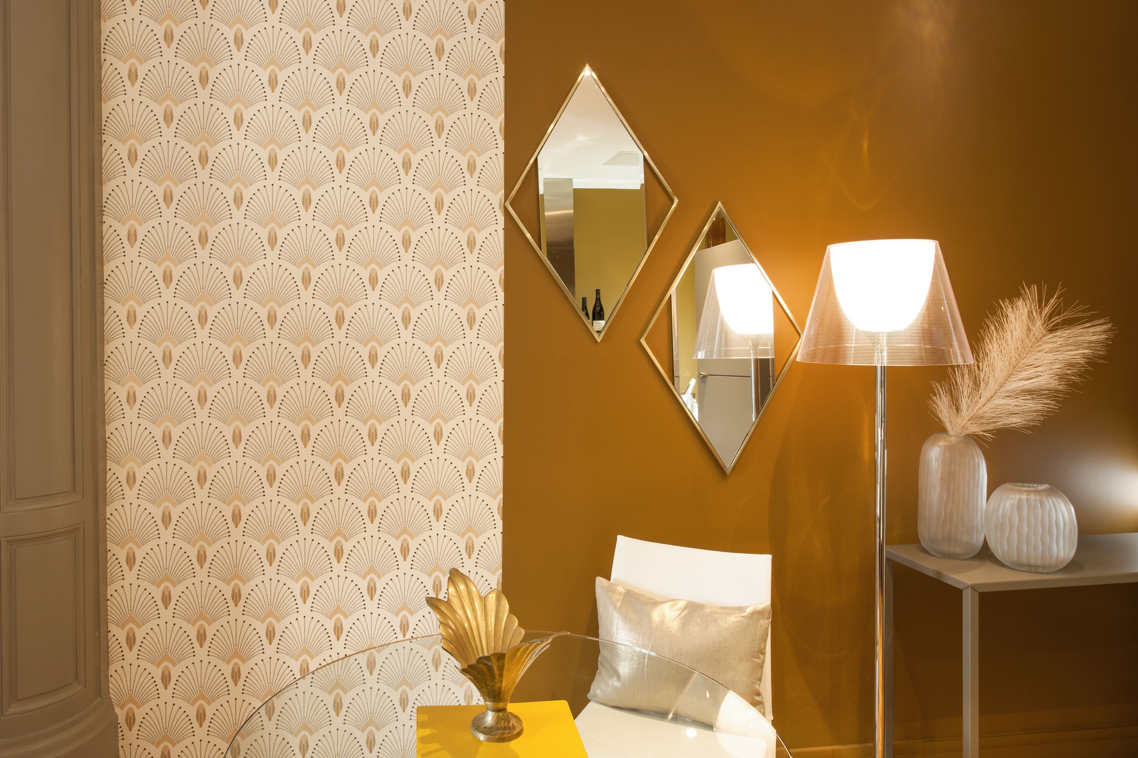 Una habitación con papel tapiz estampado, una cama con ropa blanca, dos espejos montados en la pared, una lámpara de pie y una mesita con adornos.
