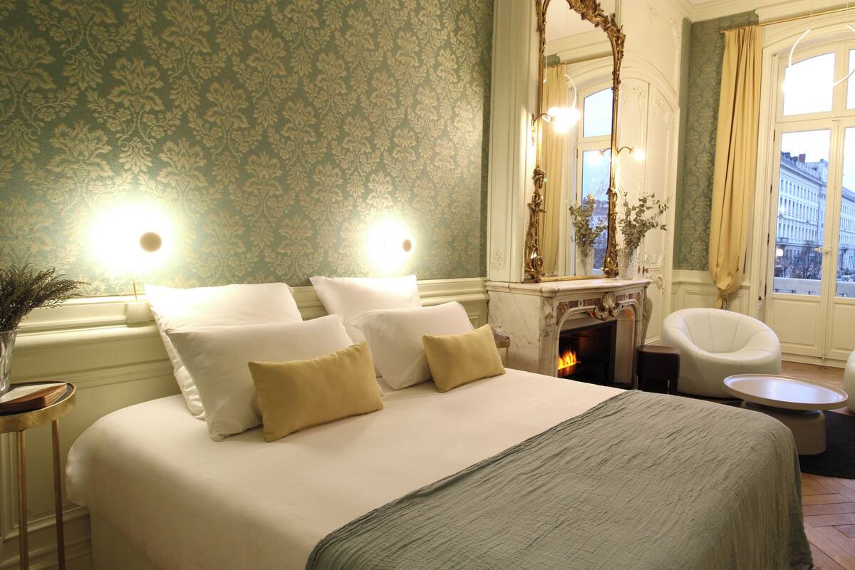 A hotel room with a bed, a fireplace, a chair, and a view of the city.