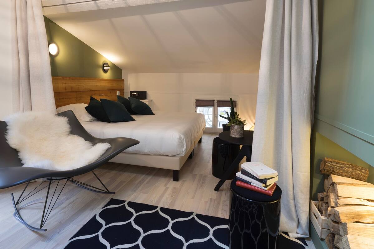 A modern bedroom with a bed, a rocking chair, a bedside table with books, and a small potted plant.