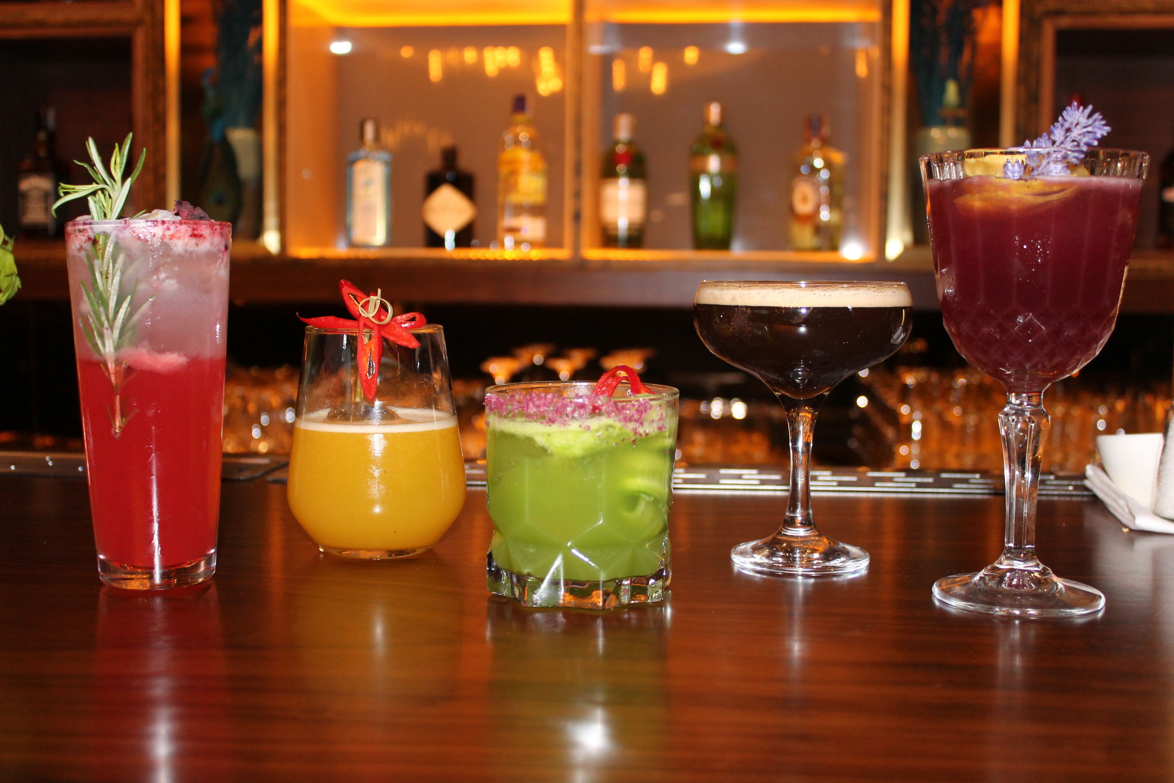 A bar with various drinks, including a red cocktail, a yellow drink, a green drink, a dark drink, and a purple drink, each garnished differently.