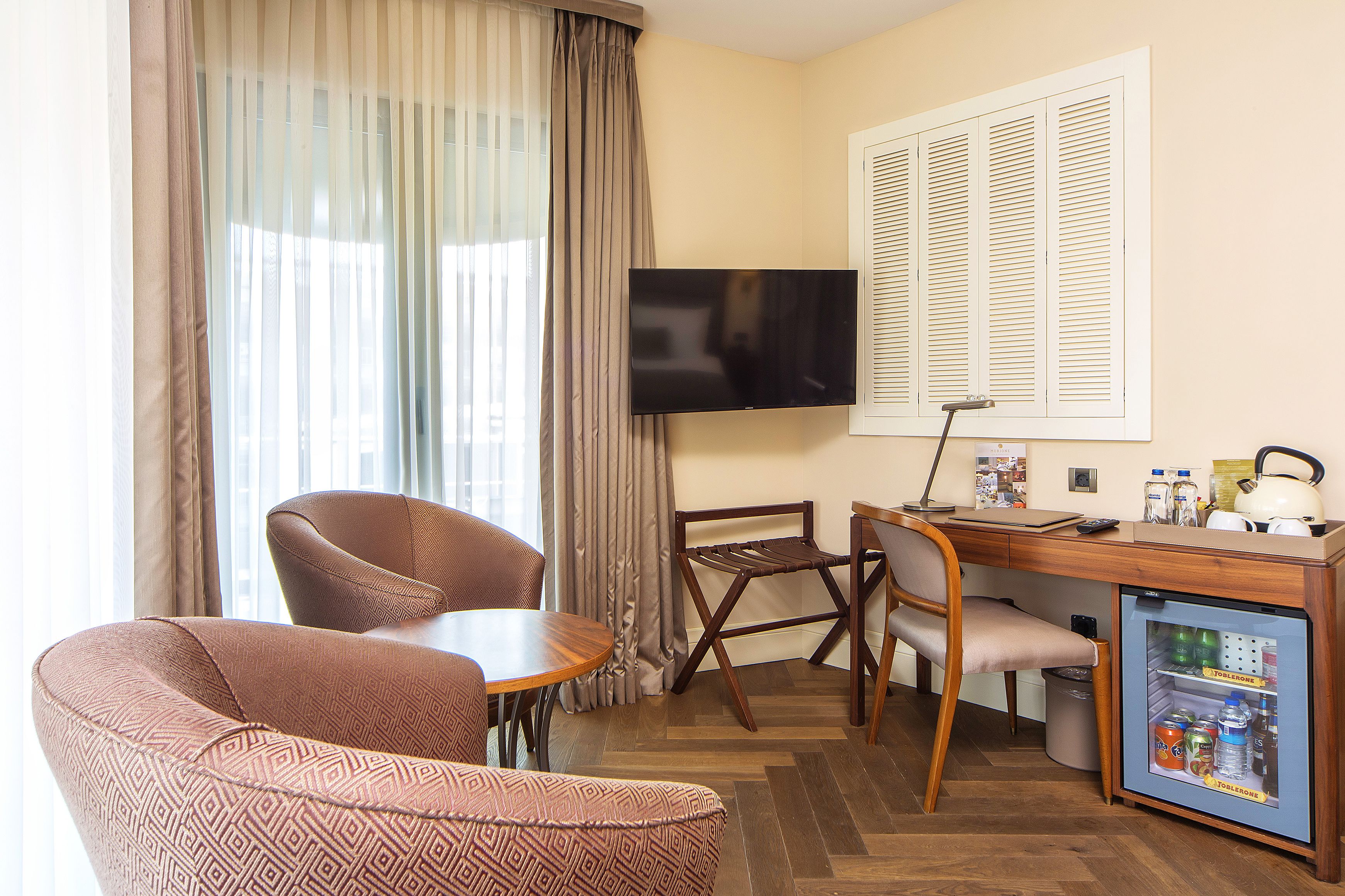A compact hotel room with a wooden desk, two chairs, a television, a mini-fridge, and a window with curtains.