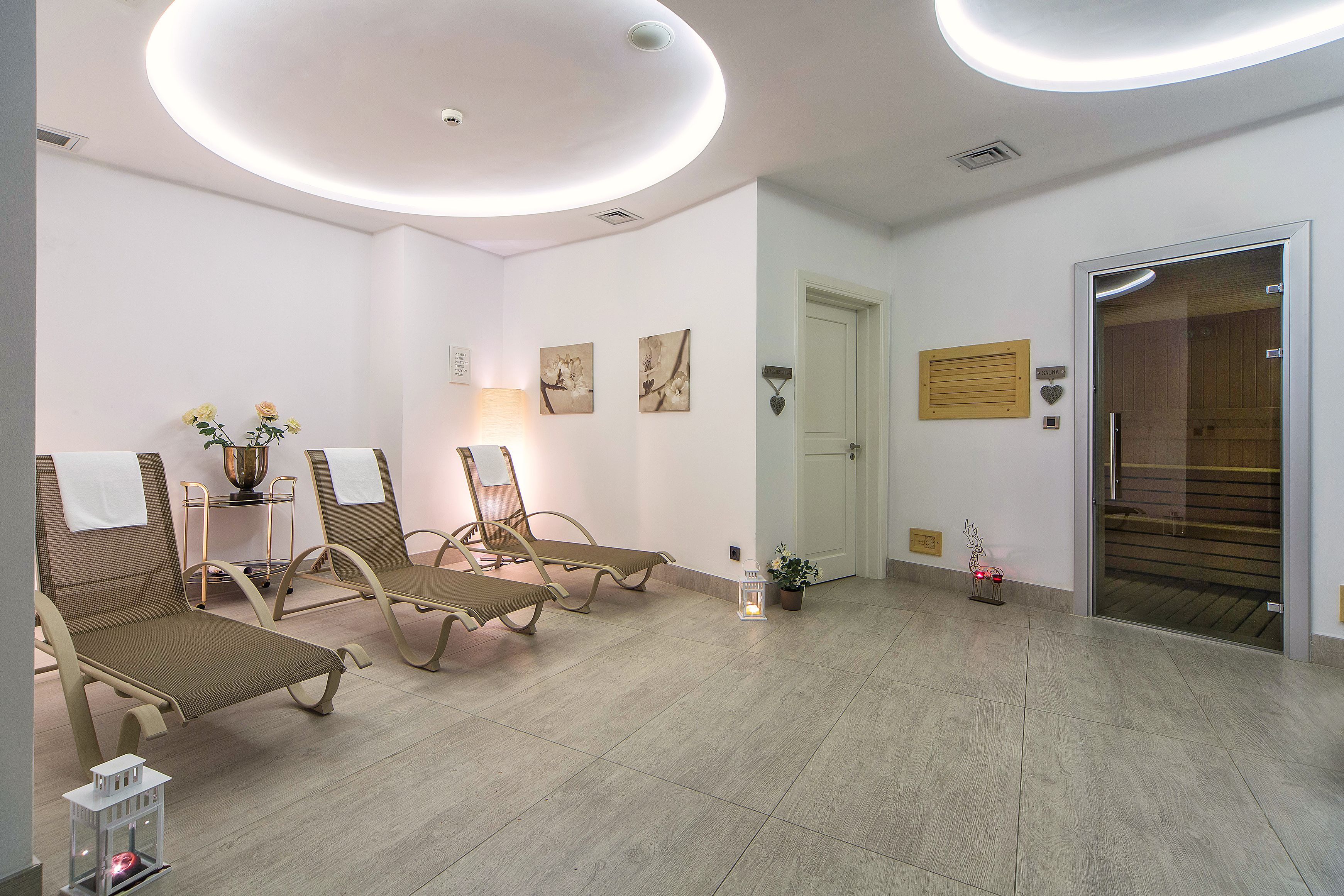 Ein Spa-Raum mit Liegesesseln, einer Massageliege, einem kleinen Tisch mit Blumenvase und einer Tür zum Sauna-Bereich.