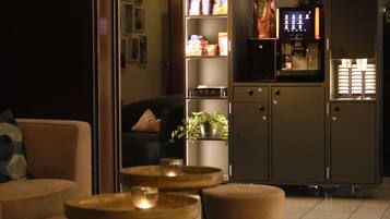 Una sala de estar moderna con un sofá, pufs, una mesa de centro y una cocina integrada que incluye un refrigerador, estantes y una cafetera.