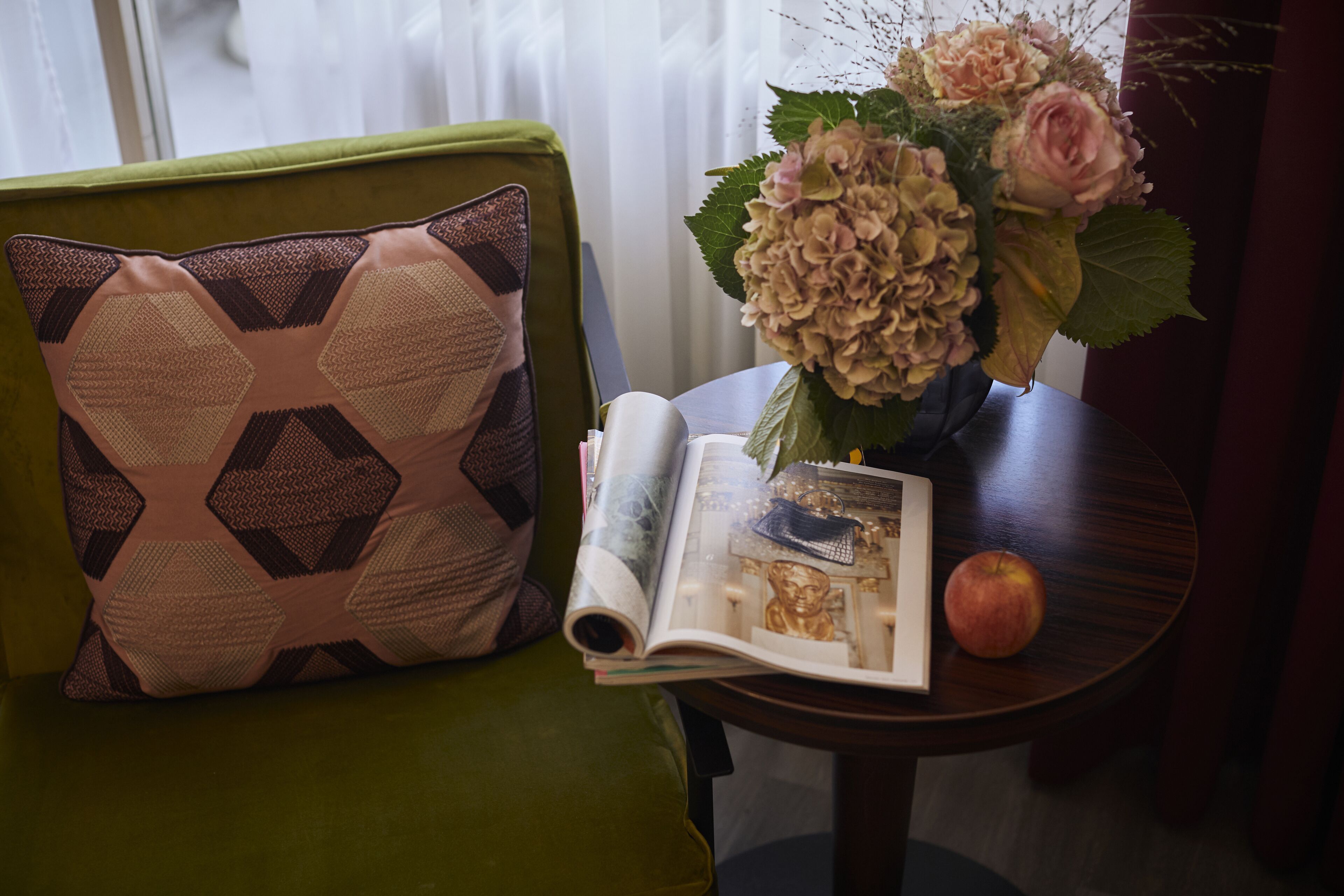 Un sillón verde con un cojín estampado, una mesita redonda con un libro, un florero con flores y una manzana.