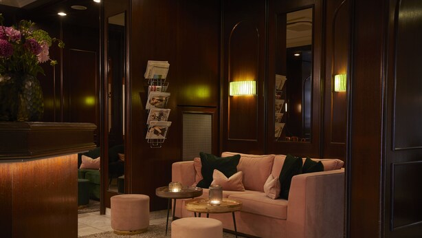 A modern living room with a pink sofa, round coffee table, and ottoman, a wooden reception desk, and a wall-mounted lamp. - Hotel Metropol by Maier Privathotels (Munich)