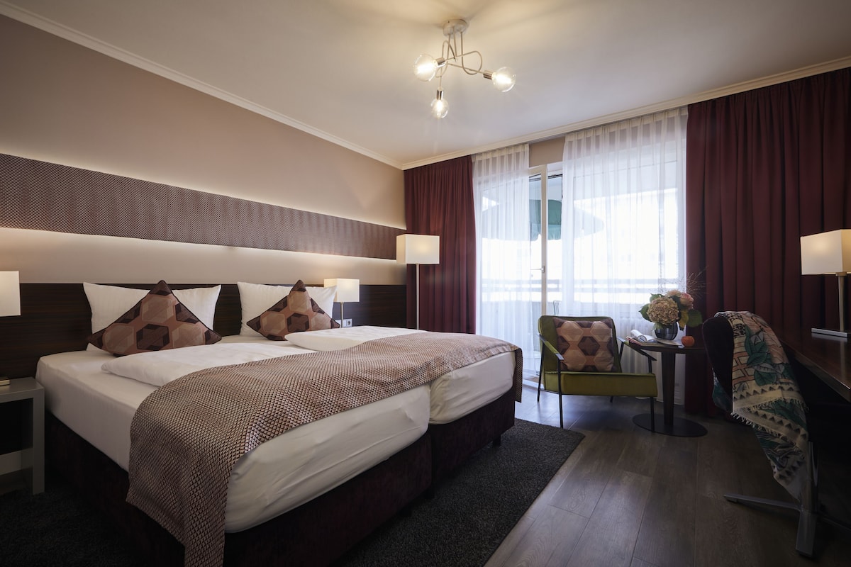 A hotel room with two beds, a desk, a chair, and a large window with curtains.