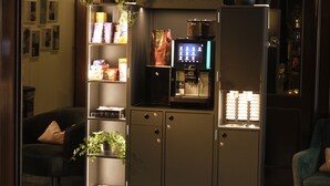 A modern lobby with a coffee machine, seating area, and a plant. - Hotel Metropol by Maier Privathotels (Munich)