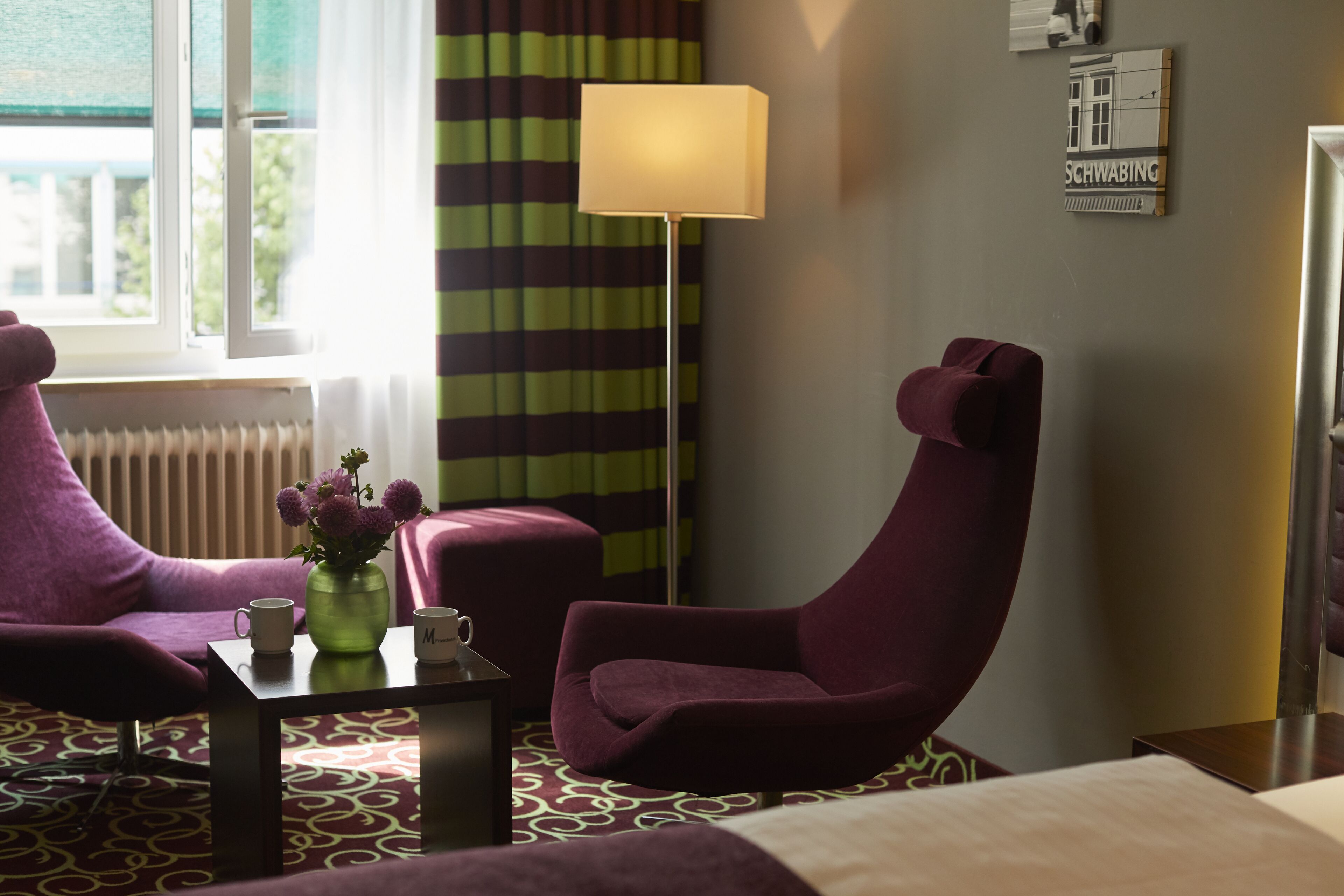 A hotel room with a purple armchair, a small table with a vase and two mugs, a floor lamp, and a view of the outdoors through a window with striped curtains.