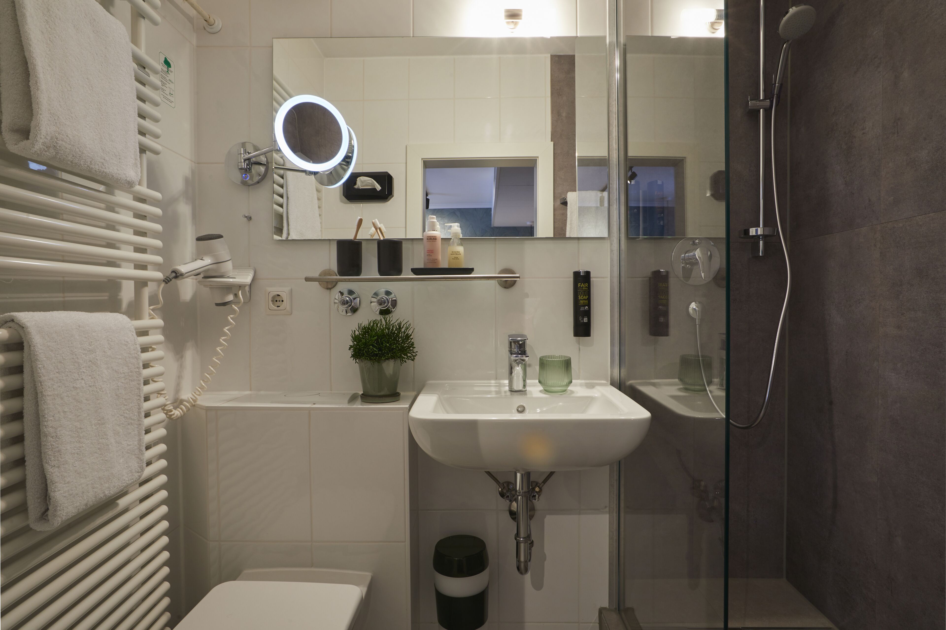Une salle de bain moderne avec un lavabo blanc, une douche à parois de verre, un miroir doté d’un anneau lumineux et un porte-serviettes.