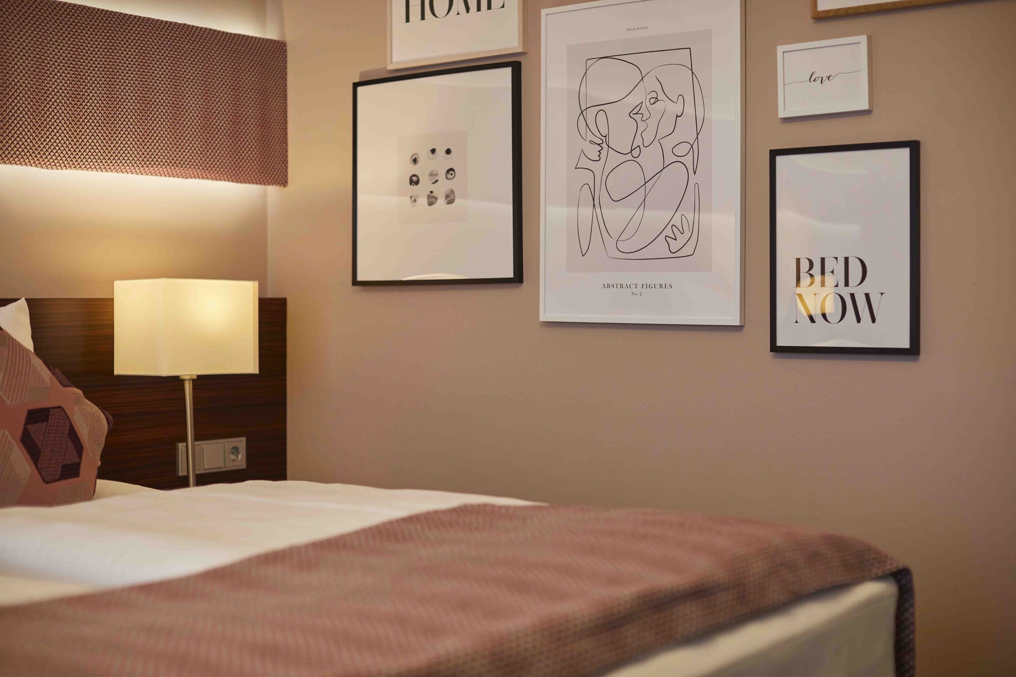 A hotel room with a bed, a wall-mounted lamp, and several framed artworks including 'HOME', 'ABSTRACT FIGURES', 'love', and 'BED NOW'.