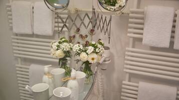Un baño con repisa de vidrio que contiene artículos de aseo, un espejo y un florero con flores.