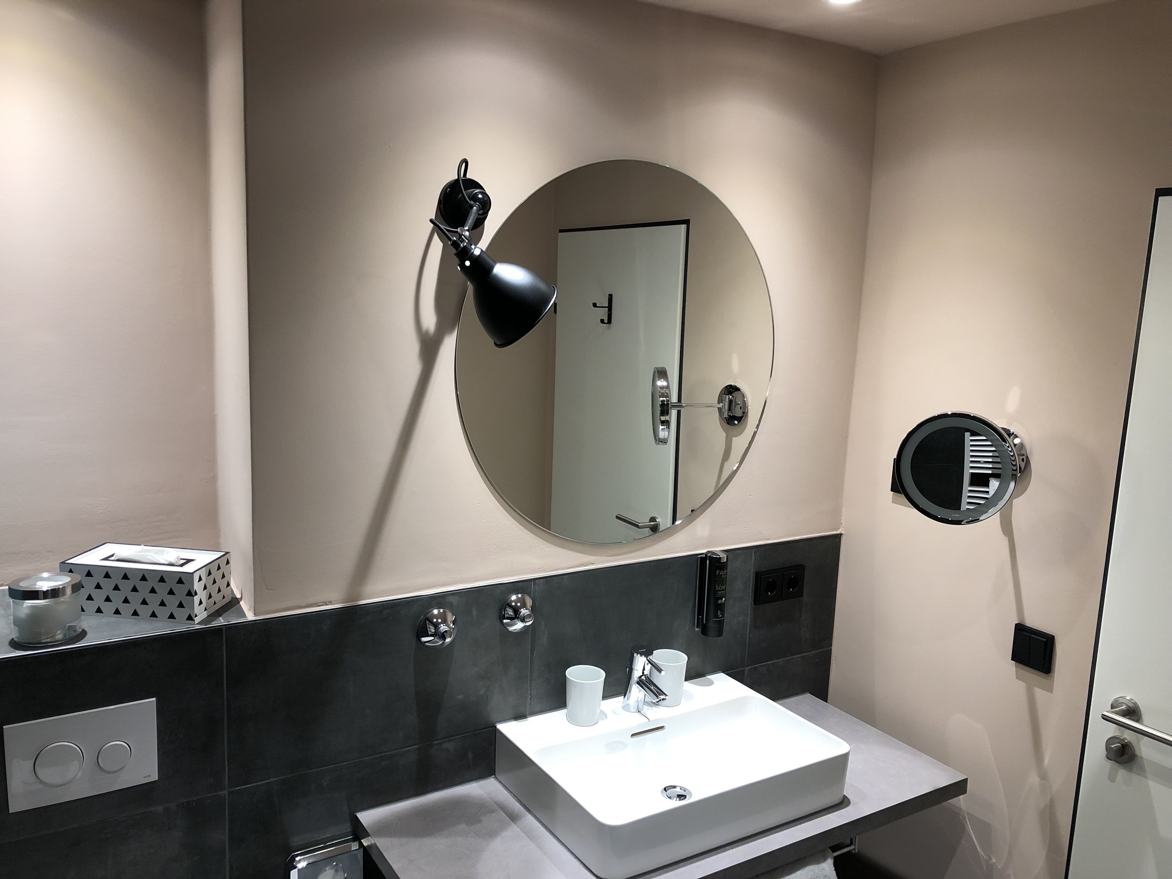 Un baño moderno con un lavabo blanco, un espejo redondo, un secador de pelo y un perchero para toallas.