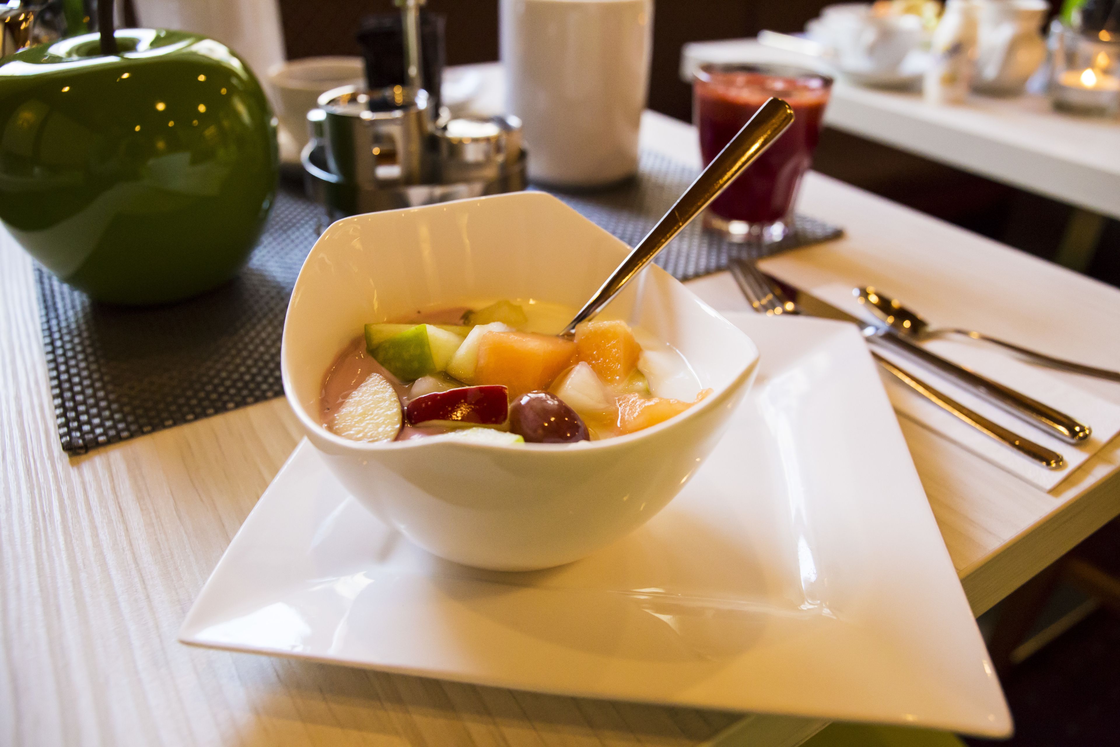 A bowl of fruit salad on a white plate with a spoon, placed on a table with a placemat.