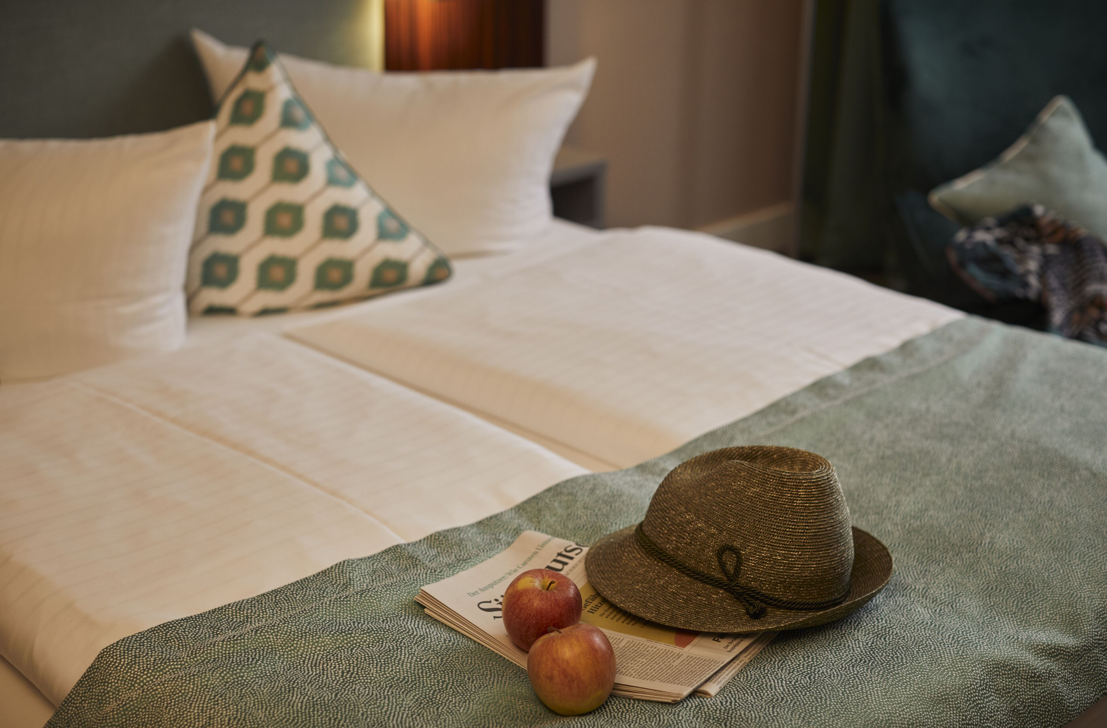 Una cama bien tendida con una colcha verde, dos manzanas y un sombrero sobre la cama.