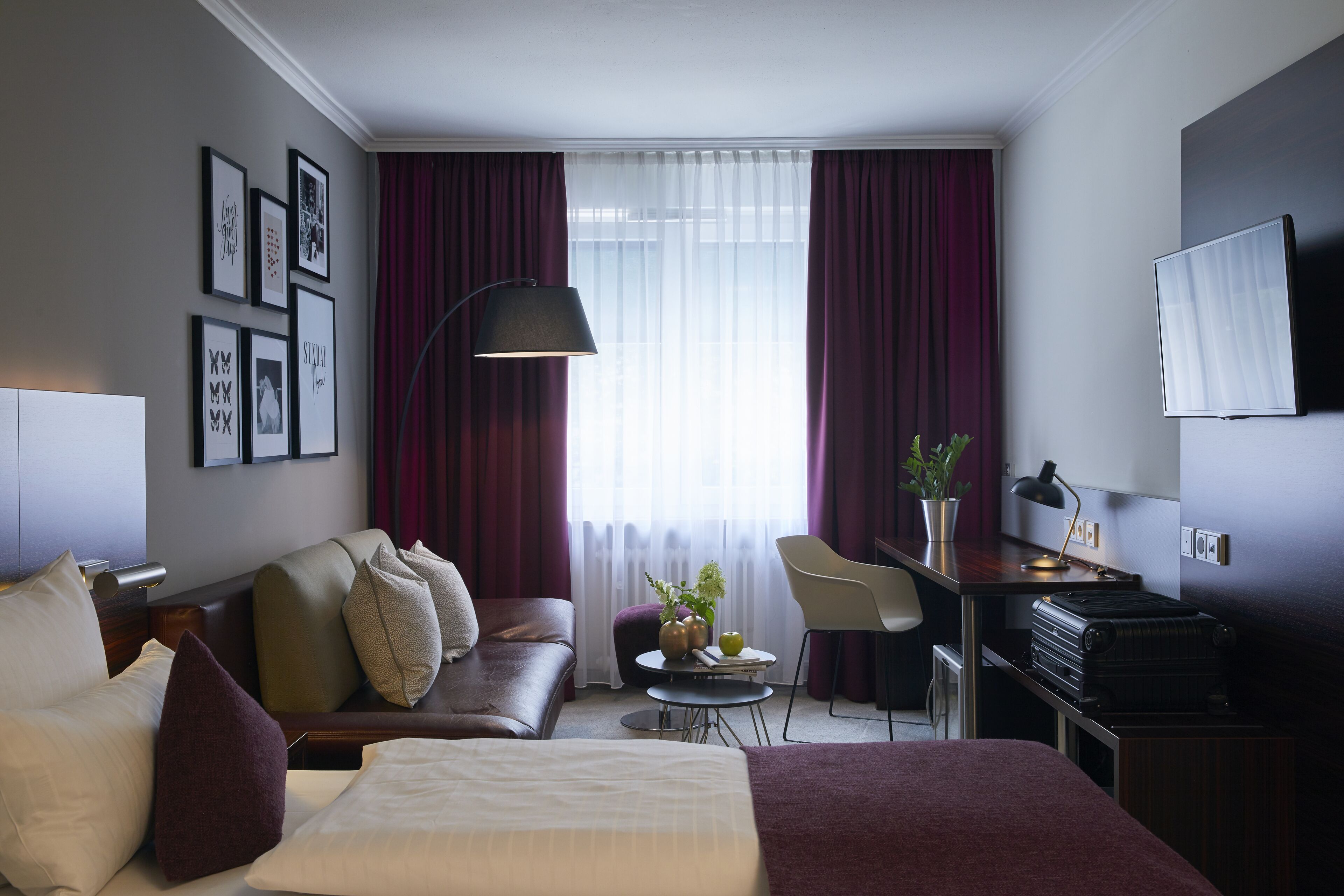 Habitación de hotel con cama, sofá, escritorio con silla, televisor y ventana con cortinas.