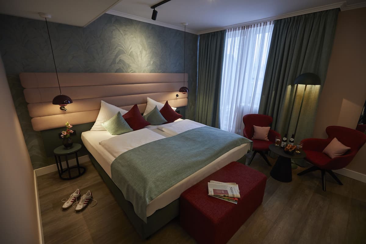A modern hotel room with a large bed, red armchairs, a small table, and a nightstand.