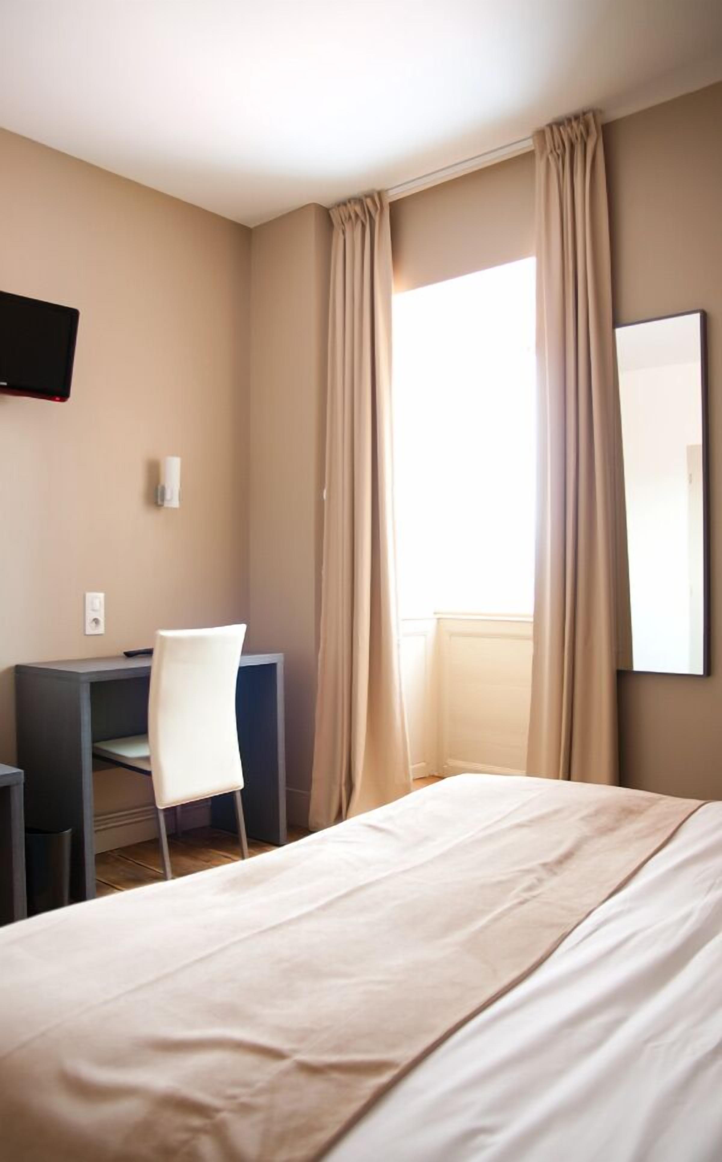 A hotel room with a bed, a desk, a chair, a TV mounted on the wall, and a window with curtains.
