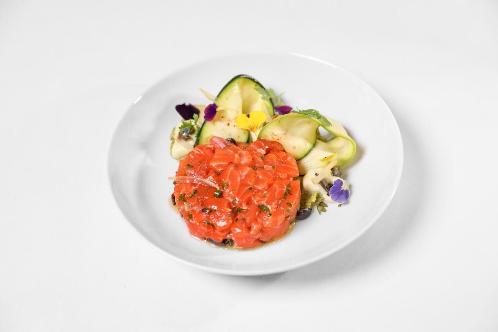 A plate with a salmon tartare centerpiece, surrounded by sliced cucumbers and a mixed greens salad with purple flowers.