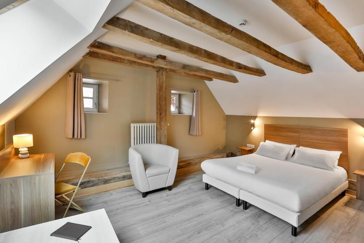 A modern bedroom with a sloped ceiling, exposed wooden beams, a bed, a chair, a desk, and a lamp.