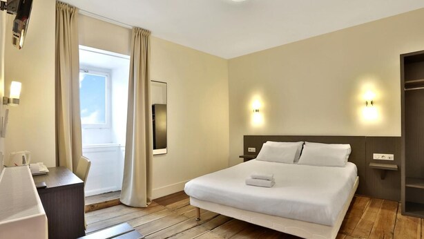 A hotel room with a large bed, a desk, a chair, a window with curtains, and a wall-mounted TV. - Auberge Dupuytren (Pierre-Buffiere)