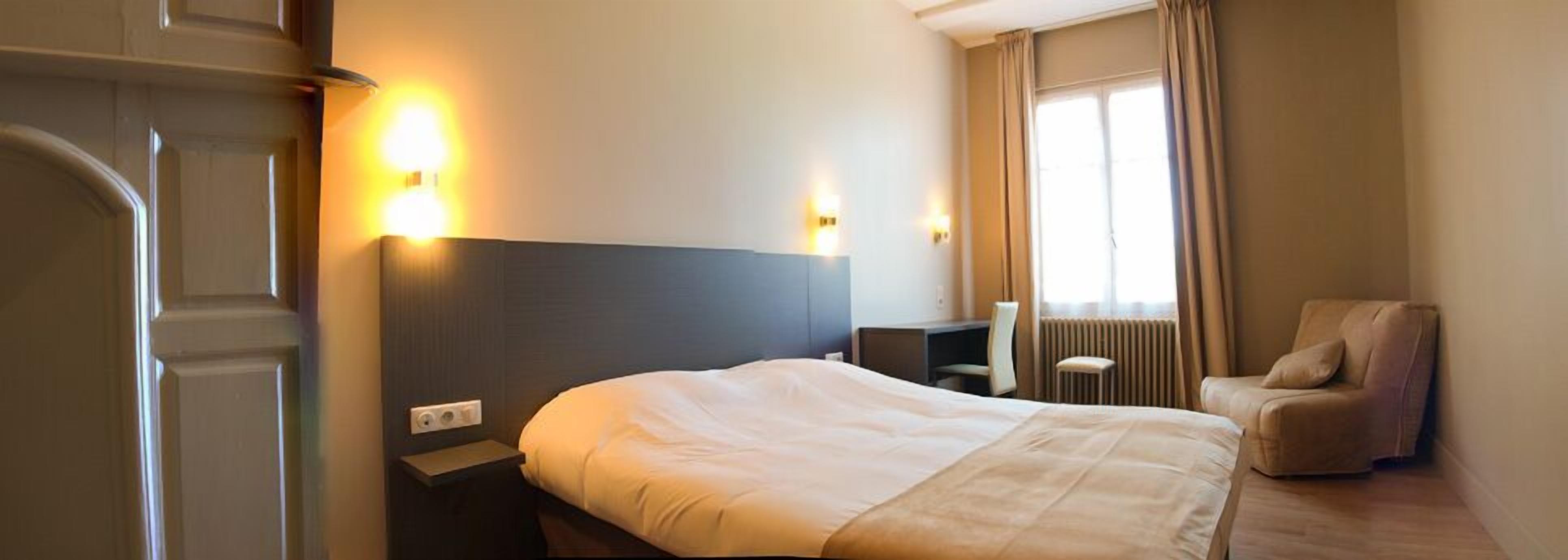 A hotel room with a bed, bedside table, lamp, chair, and a window with curtains.