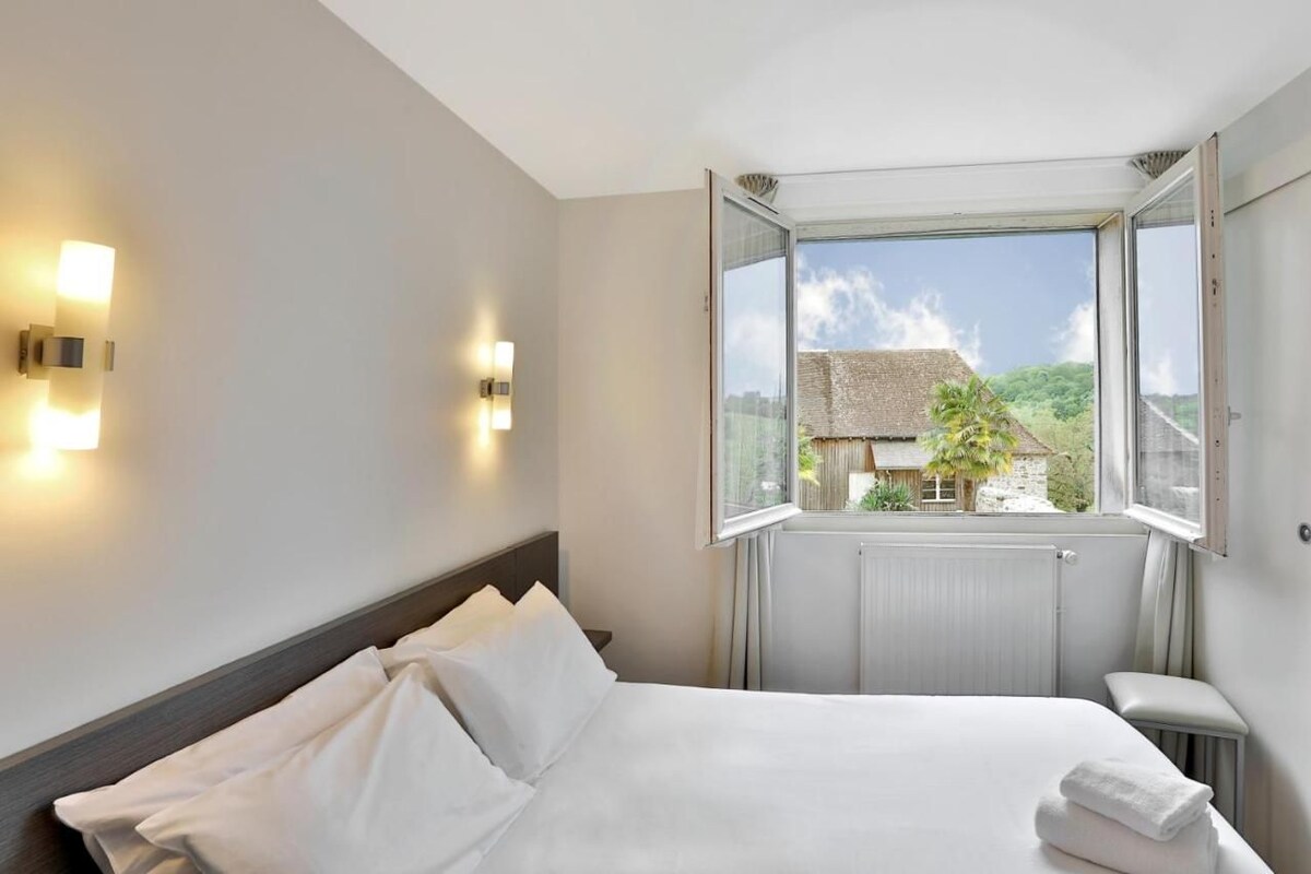 A hotel room with a bed, two wall-mounted lights, a window with an open view of a house and greenery.