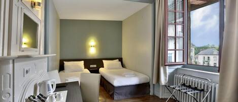A hotel room with two beds, a desk with a chair, a radiator, and a window with a view of buildings.