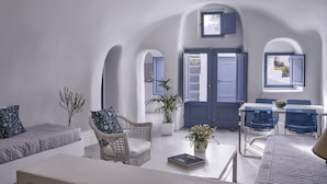 A modern living room with a white sofa, a wicker chair, a dining table with blue chairs, and a view through an arched doorway. - Fava Eco Suites (Santorini)