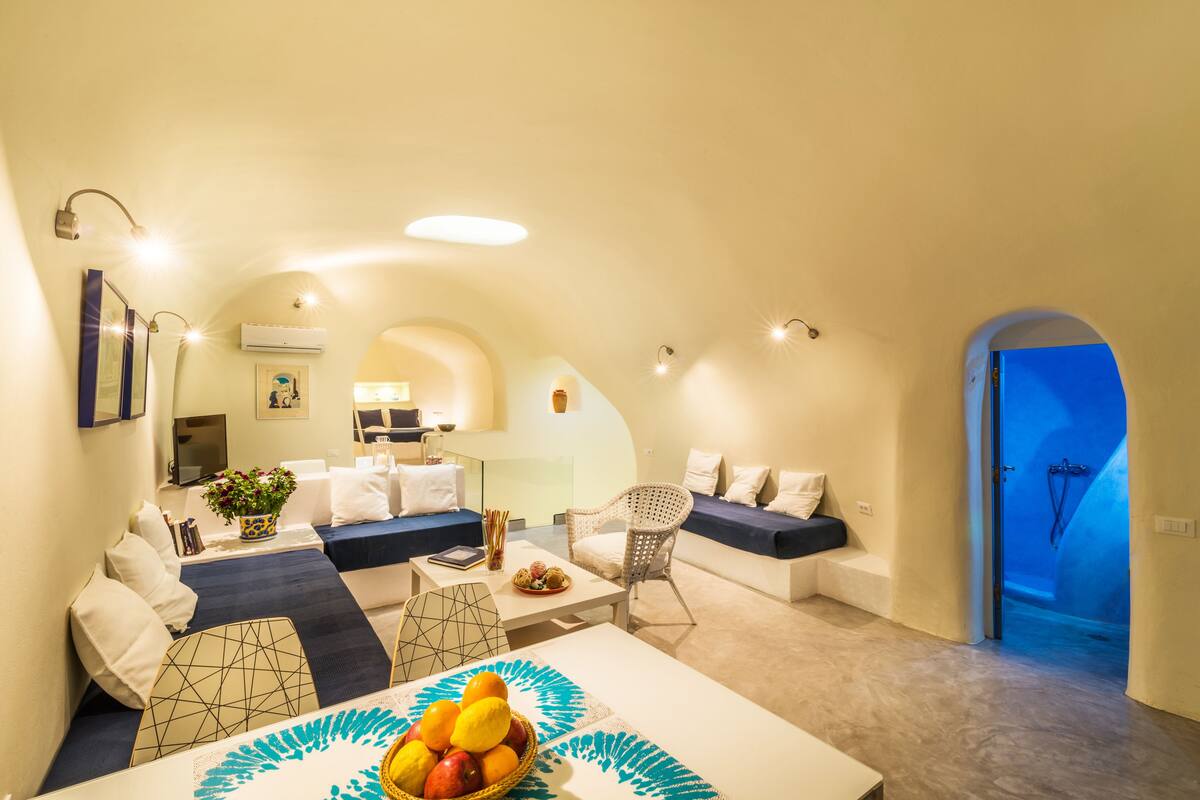 A modern, well-lit living area with a sofa, a dining table with a fruit bowl, and a blue door leading to a bathroom.