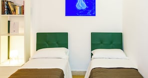 Due letti singoli con poggiatesta verdi, una parete bianca, una libreria con libri e un quadro astratto blu.