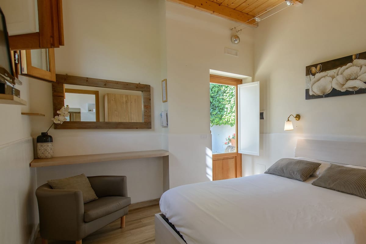 A bedroom with a large bed, a wooden headboard, a bedside table with a lamp, a small sofa, and a wall-mounted artwork.