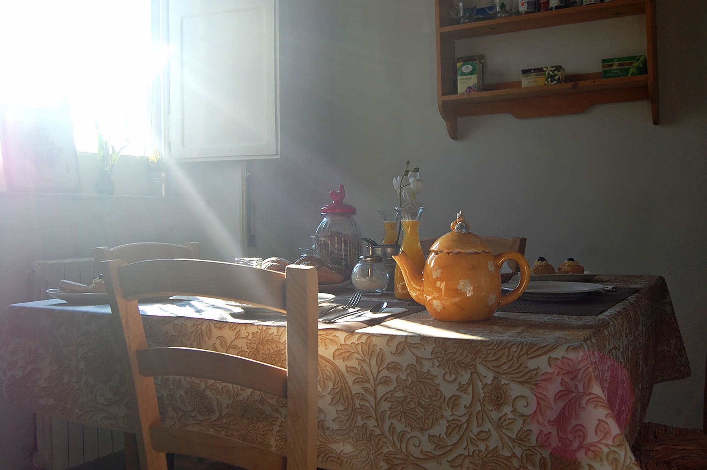 Una mesa de comedor con mantel floreado, una tetera y una jarrón. Encima de la mesa hay una estantería con varios objetos.