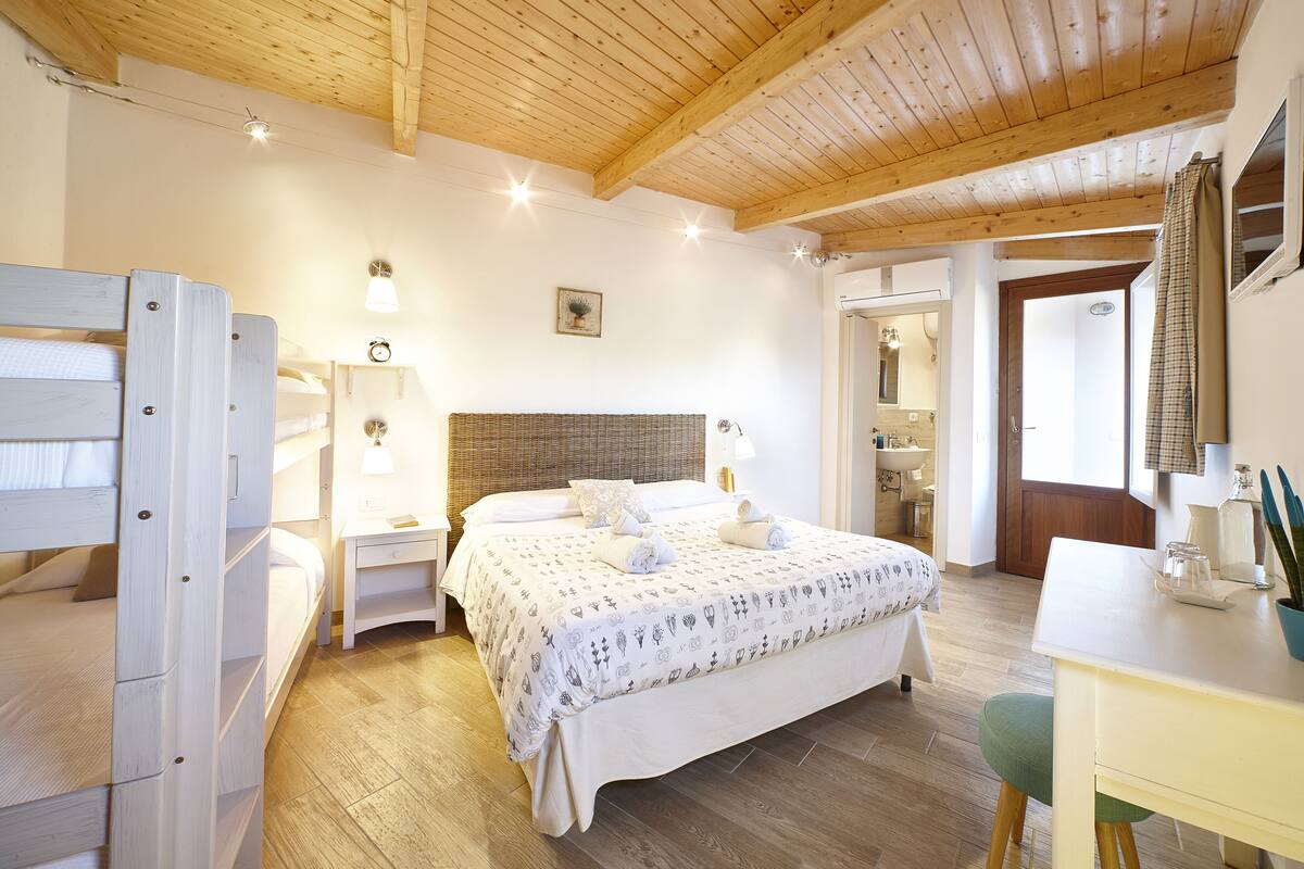 A bedroom with a wooden ceiling, a bed with a patterned bedspread, bedside tables with lamps, and a desk with a plant.