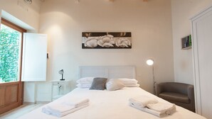 Una cama bien hecha con sábanas y toallas blancas, una mesita de noche con lámpara, una obra de arte en la pared y una ventana con vistas a la vegetación.