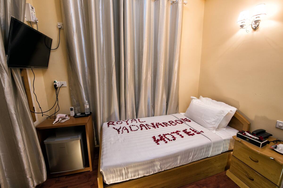 Una habitación de hotel con una cama, una mesita de noche de madera, un televisor montado en la pared y una cortina.